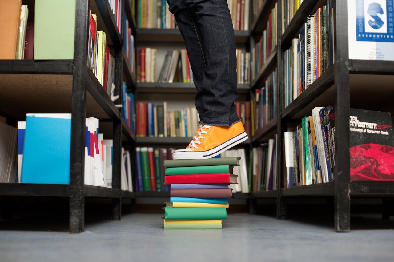 A person standing on a colorful stack of books in a library aisle.